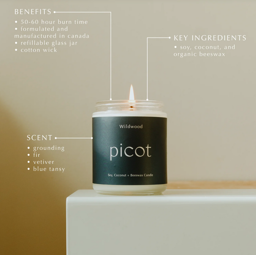 Lit "picot Wildwood" candle on white surface. Dark label lists benefits (long burn, Canadian-made, refillable, cotton wick), scents (grounding, fir, vetiver, blue tansy), and ingredients (soy, coconut, beeswax).