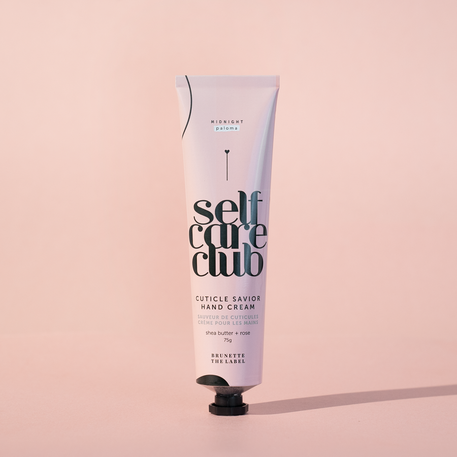 A pale pink tube of "Midnight Paloma Self Care Club Cuticle Savior Hand Cream" with text in French and English, featuring "shea butter + rose," is standing upright on a matching pale pink background. The bottom of the tube indicates "BRUNETTE THE LABEL."