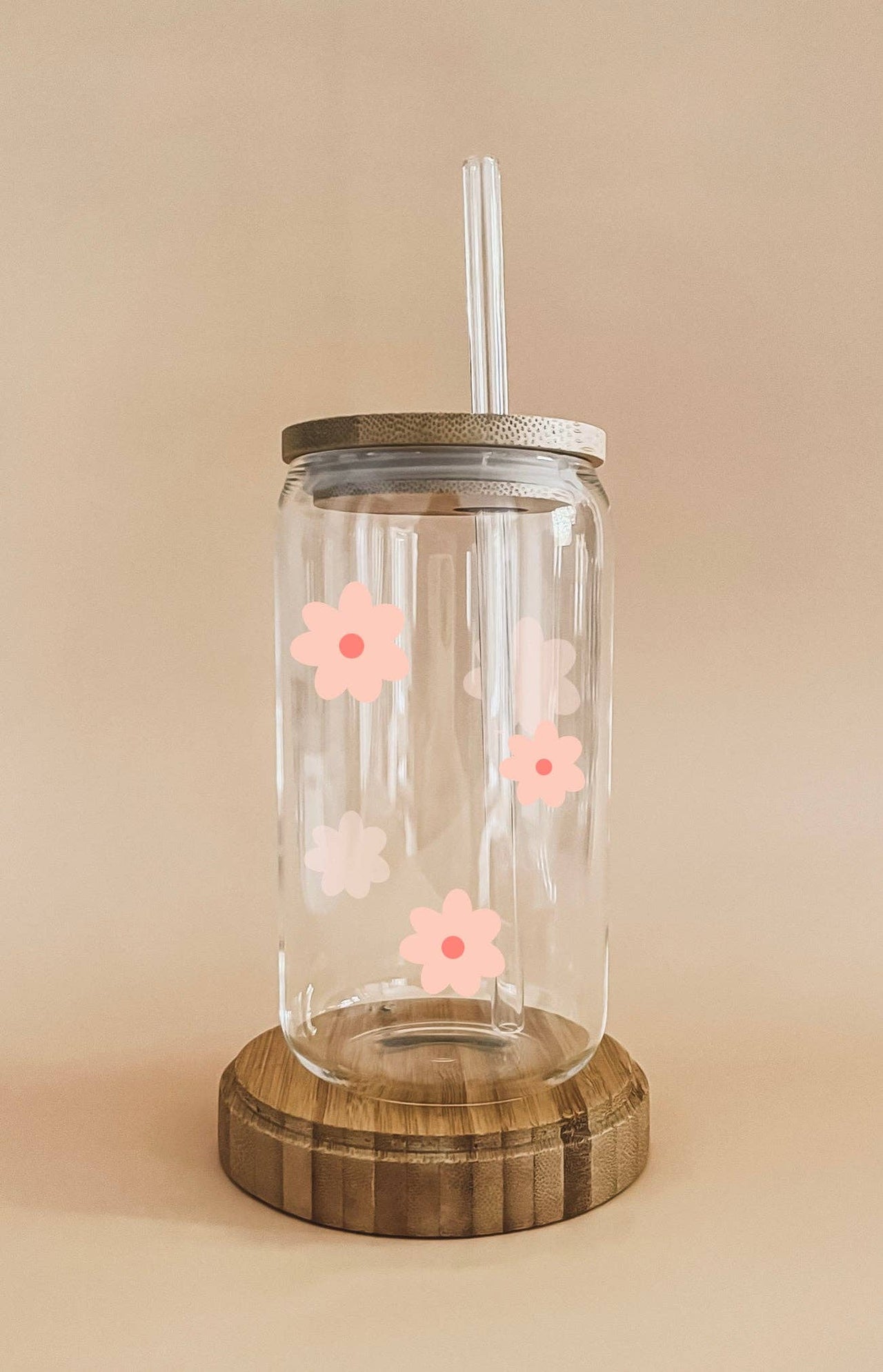 A clear glass reusable cup with a bamboo lid and straw is decorated with pink and white flower decals and rests on a round wooden coaster.