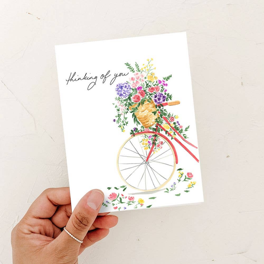 A hand holding a watercolor "Thinking of You" card featuring a vintage red bicycle with a basket full of vibrant flowers against a white textured background.