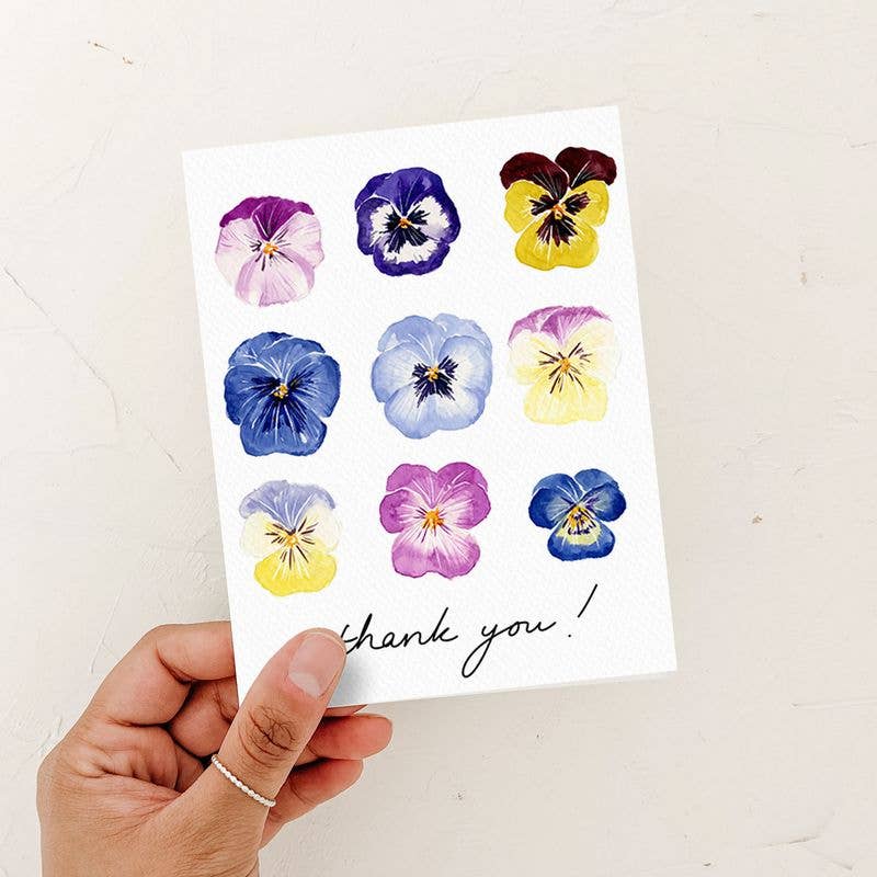 A person's hand holding a watercolor "Thank You!" card featuring a grid of nine colorful pansies against a rustic white background.