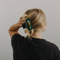 Thumbnail for From behind, a person with blonde hair is seen securing their messy bun with a dark green, rectangular hair claw clip.