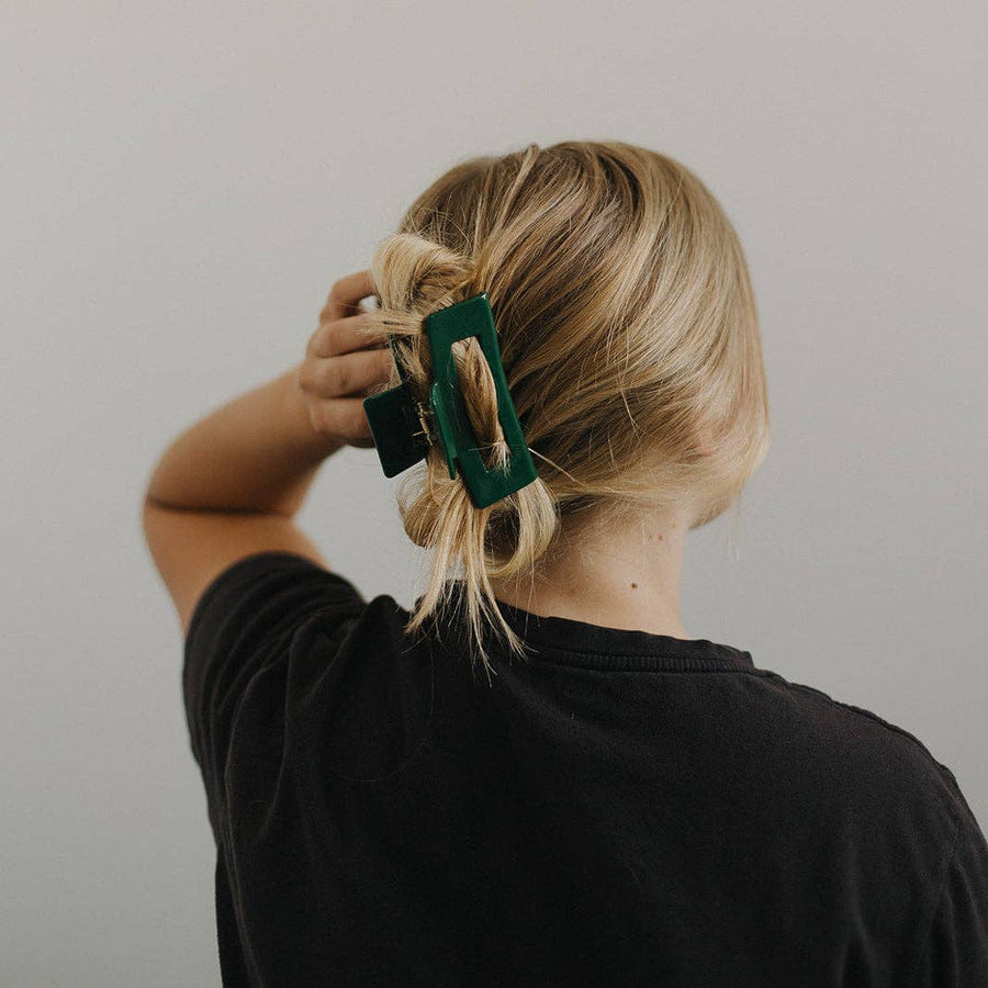 From behind, a person with blonde hair is seen securing their messy bun with a dark green, rectangular hair claw clip.