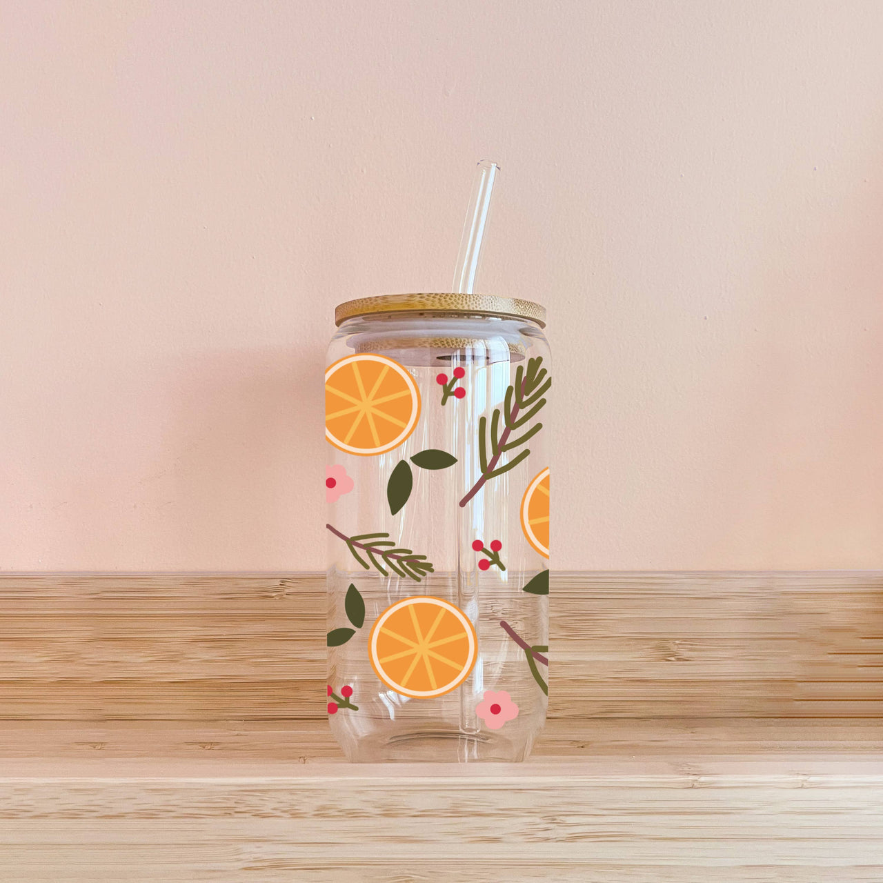 Clear glass cup with a bamboo lid and glass straw, decorated with a repeating pattern of orange slices, evergreen branches, and small pink flowers.