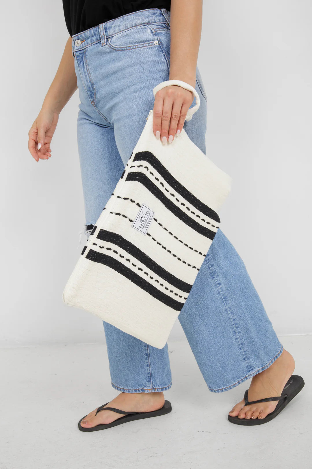 A side-view of a person walking while holding the large Ecru/Black striped Mercer bag by its wrist strap. The bag hangs lower and appears significantly more spacious than the small version, reaching from the waist down to the mid-thigh.