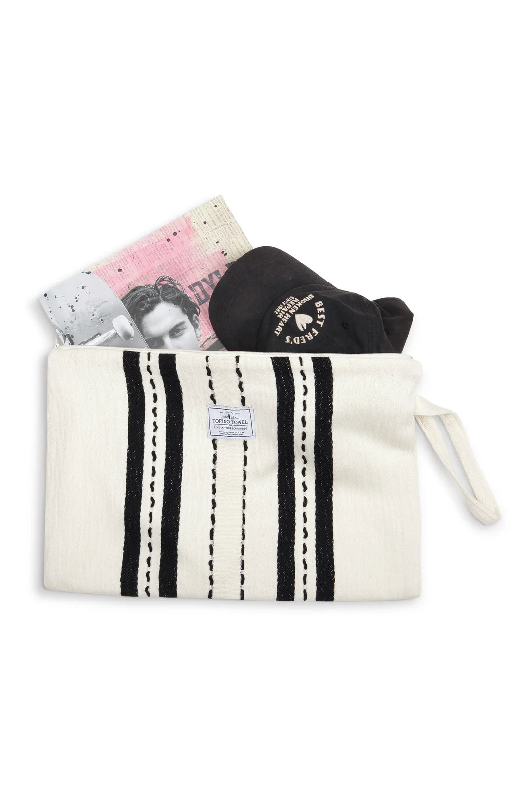A top-down flat lay of the large Ecru/Black striped Mercer bag. Inside the open zipper are a black baseball cap and a large magazine, showing the bag's ability to hold oversized travel or adventure essentials.